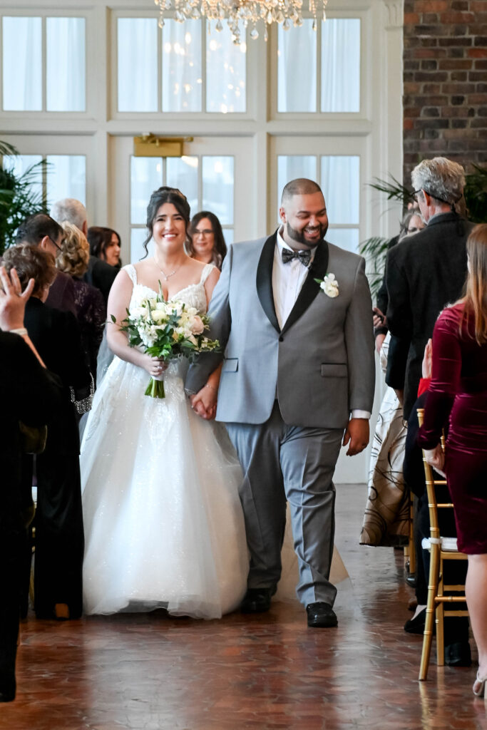 Bride and groom walking hand in hand down the aisle, smiling joyfully as guests celebrate their wedding at The Virginia Cavalier.