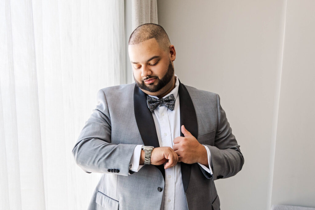 Groom adjusting his watch while standing near a window with soft natural light, preparing for his wedding day at The Marriott