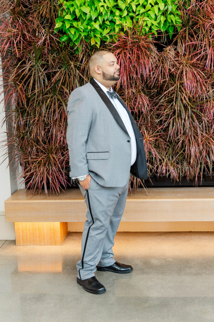 Groom standing confidently in a light gray suit with a black lapel, posing in front of a vibrant green and red plant wall at The Marriott.