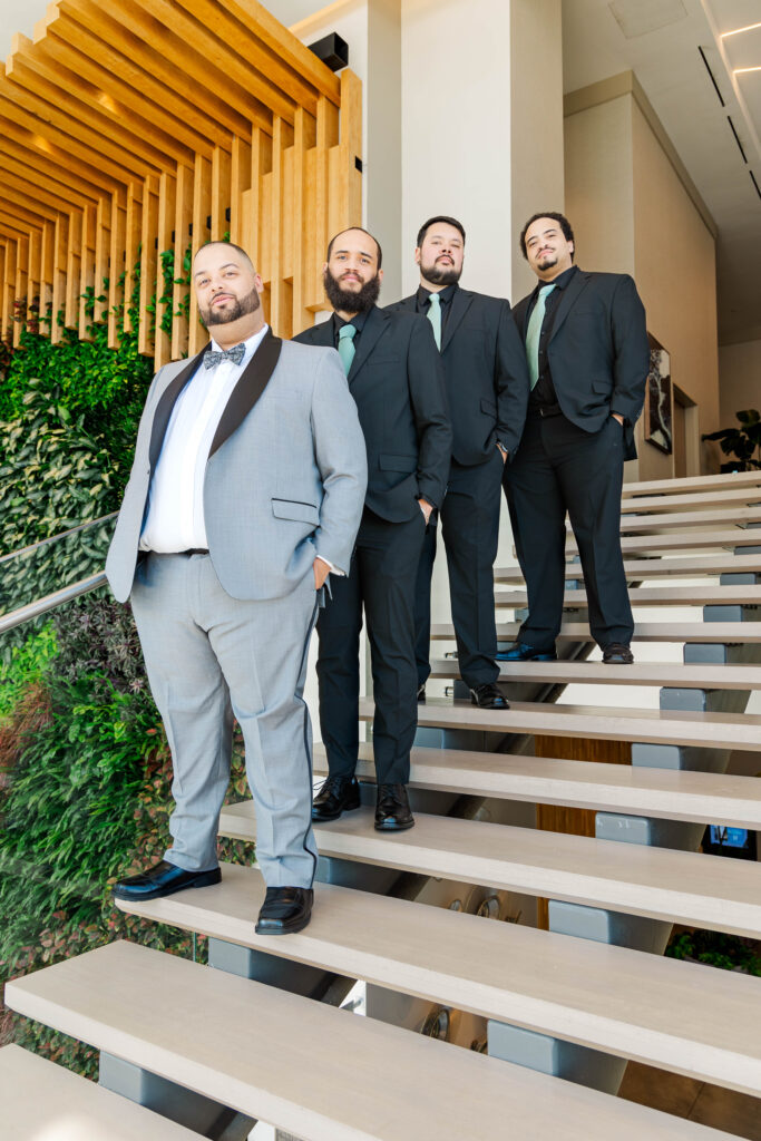 Groom and groomsmen standing confidently on modern stairs with greenery and wooden accents at The Marriott, showcasing their stylish wedding attire.