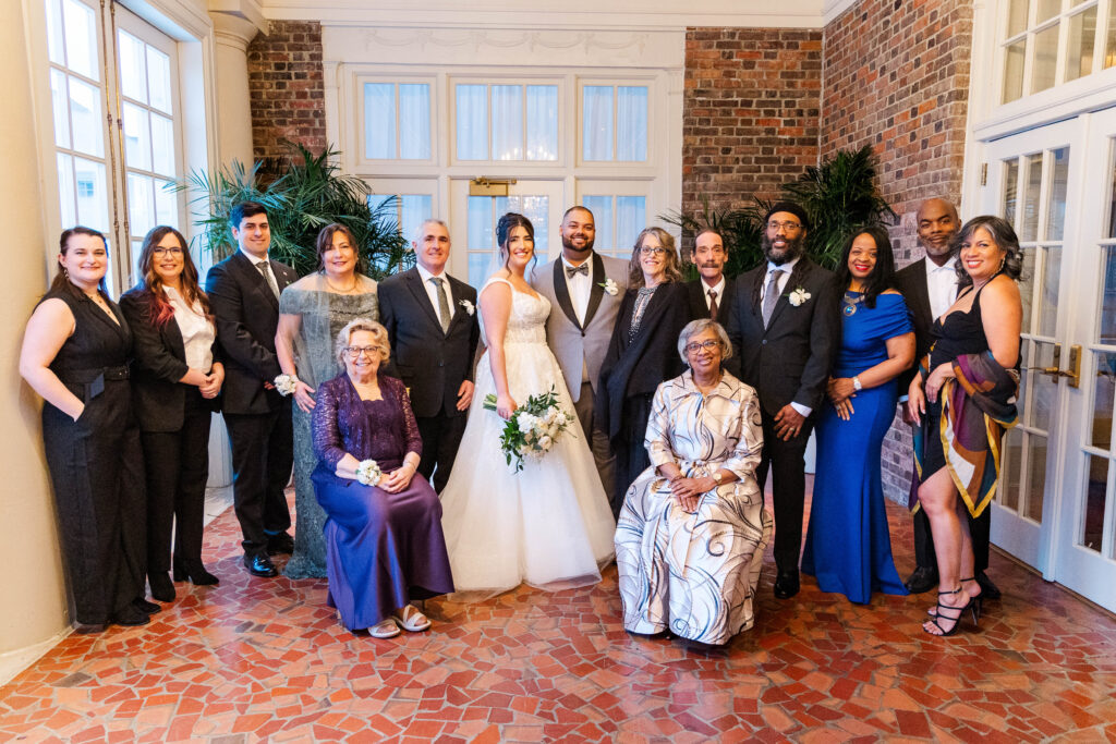 Bride and groom surrounded by their family, posing for a group photo in a beautifully lit room with brick walls and classic decor at The Virginia Cavalier.